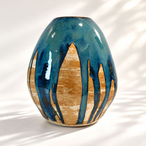 A handmade ceramic vase with a rounded, bulbous shape, featuring a glossy dark teal-blue glaze dripping over a natural tan glade, set against a white background with soft leaf shadows.