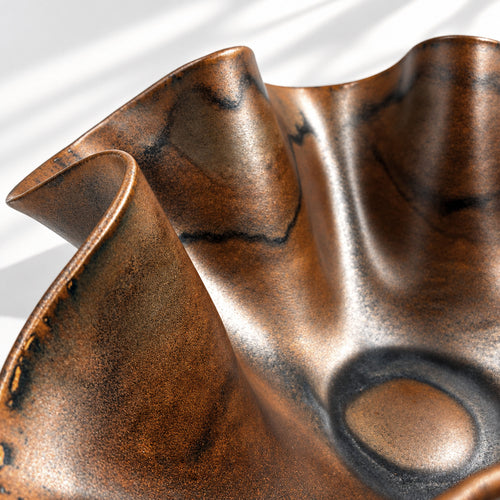 Close-up of a sculptural chocolate glaze bowl with a wavy, fluted rim and a satin finish, showing warm bronze-brown tones with darker oxidized highlights.