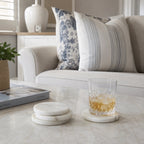 A set of round white marble coasters on a light marble coffee table, with one coaster holding a cut-glass tumbler filled with an amber drink.