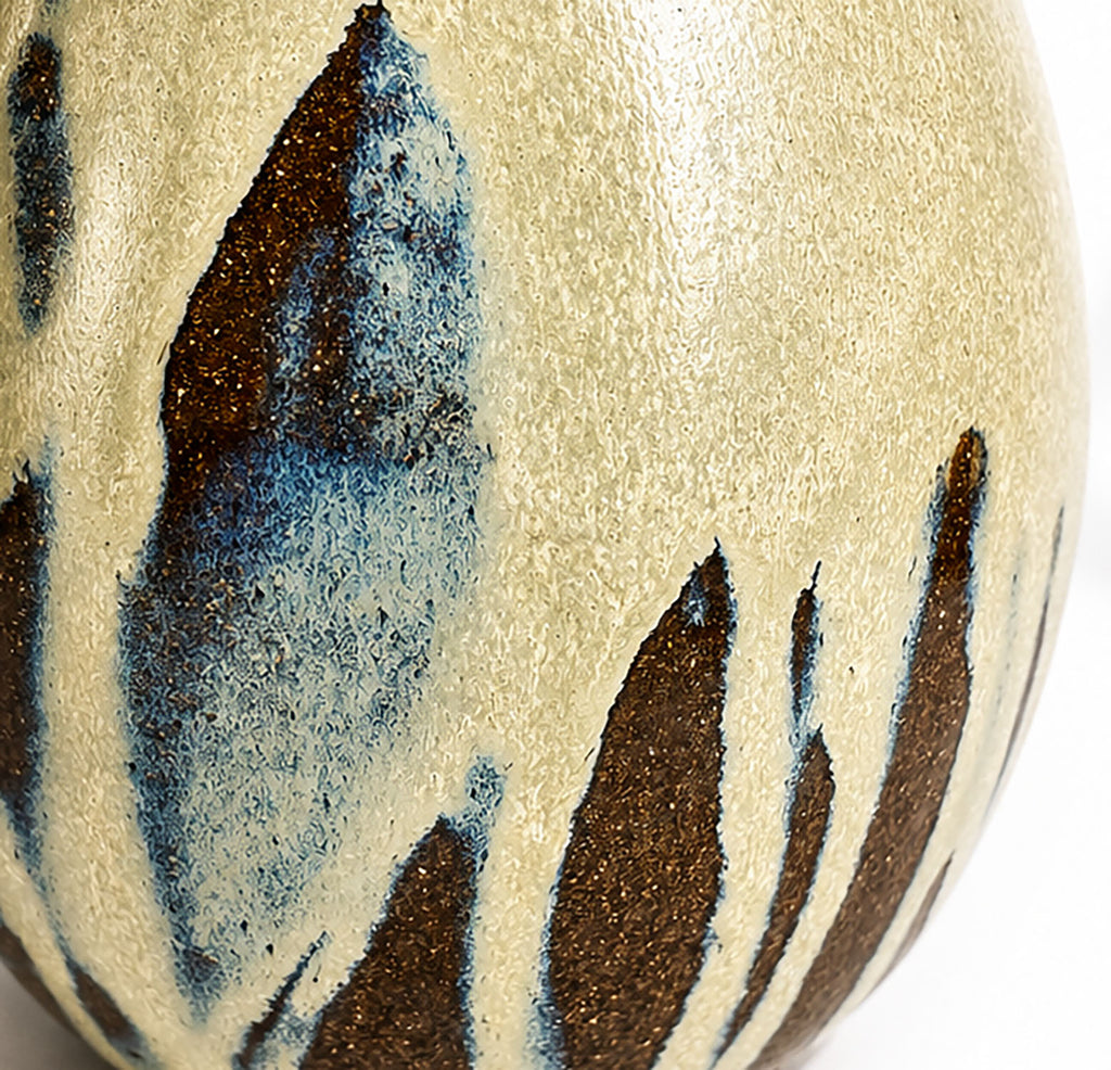Close-up of a ceramic egg vase with a textured cream-beige glaze and irregular dark brown and blue abstract brushstroke patterns.