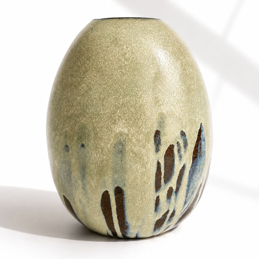 A handmade ceramic vase with a rounded, egg-shaped form, featuring a textured mint and beige glaze with dark brown and blue leaf-like accents near the base.