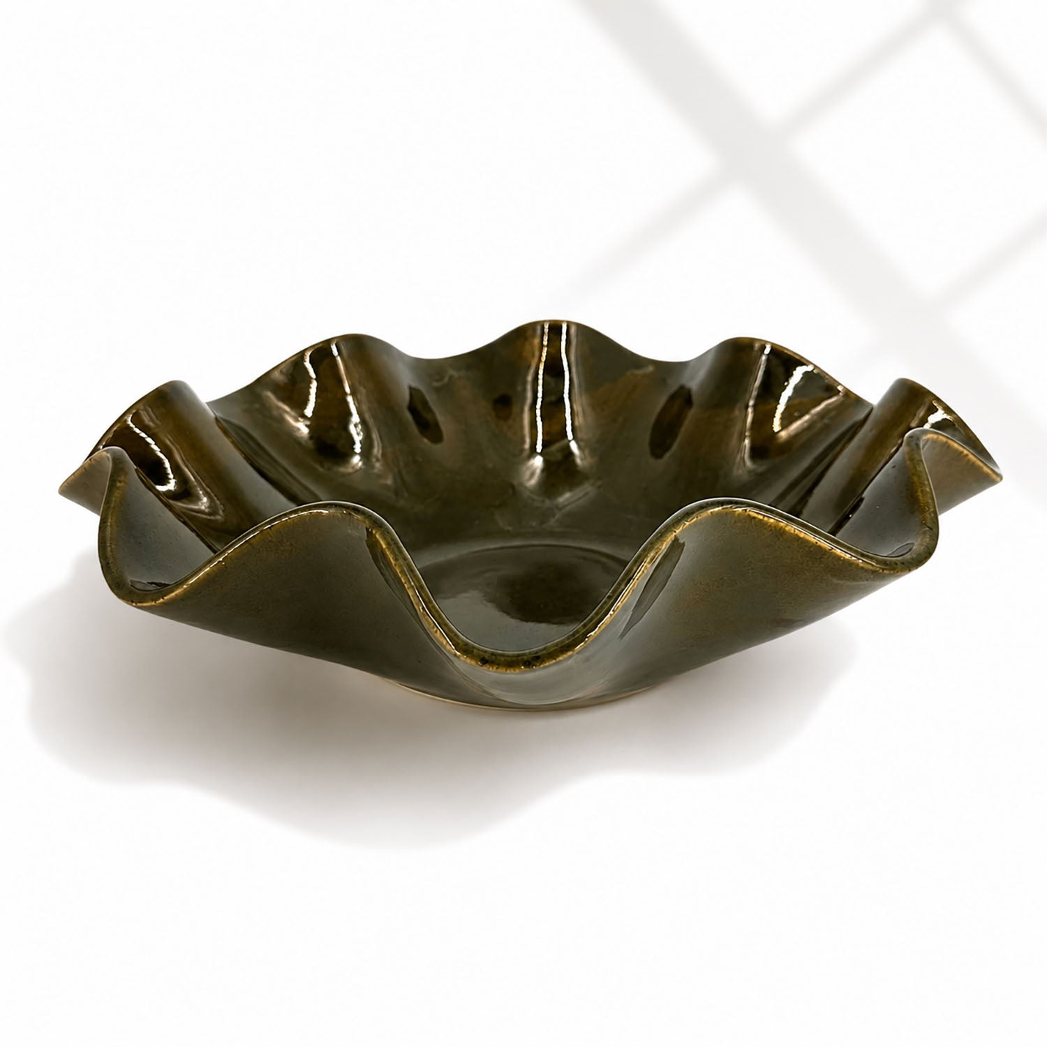 A glossy dark olive green ceramic bowl with a wavy, scalloped rim, shown on a clean white background with soft window shadow patterns.